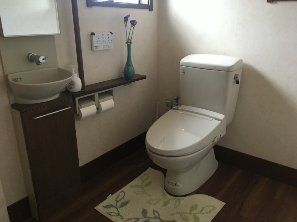 トイレの写真。一般的な家庭のトイレ風。洋式便座、右にトイレットペーパー、リモコン、手洗いがあります。